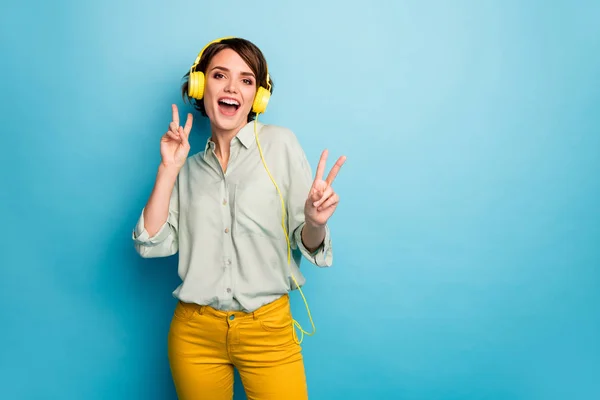 Zdjęcie wesołej pani słuchać radia fajne stylowe słuchawki disco dj pokazując v-symbole nocnych klubów klientów nosić casual zielona koszula żółte spodnie odizolowane niebieski kolor tła — Zdjęcie stockowe