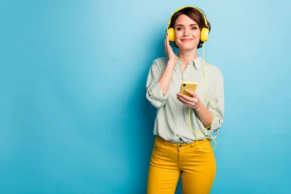 Zdjęcie śmieszne pani trzymać telefon przy użyciu nowoczesnej technologii słuchania radia fajne stylowe słuchawki nosić casual zielona koszula żółte spodnie odizolowane niebieski kolor tła — Zdjęcie stockowe