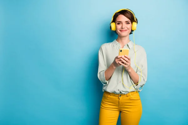 Radio rockt. Foto van mooie dame hold telefoon met behulp van moderne technologie luisteren radio oortelefoons dragen casual groen shirt gele broek geïsoleerde blauwe kleur achtergrond — Stockfoto