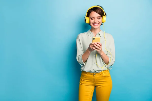 Foto van mooie dame hold telefoon met behulp van moderne technologie luisteren radio oortelefoons melodie fm dragen casual groen shirt gele broek geïsoleerde blauwe kleur achtergrond — Stockfoto
