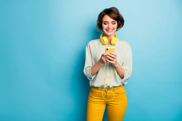 Zdjęcie ładnej młodej pani trzymać telefon przy użyciu nowoczesnej technologii włączając słuchawki piosenka nosić casual zielony koszula żółte spodnie odizolowane niebieski kolor tło — Zdjęcie stockowe