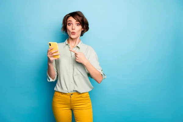 Фото красивої леді тримає телефон відкритим ротом, показуючи екран пальця, що радить низькі покупки смартфона ціна одягу звичайної зеленої сорочки жовті штани ізольовані синій колір фону — стокове фото