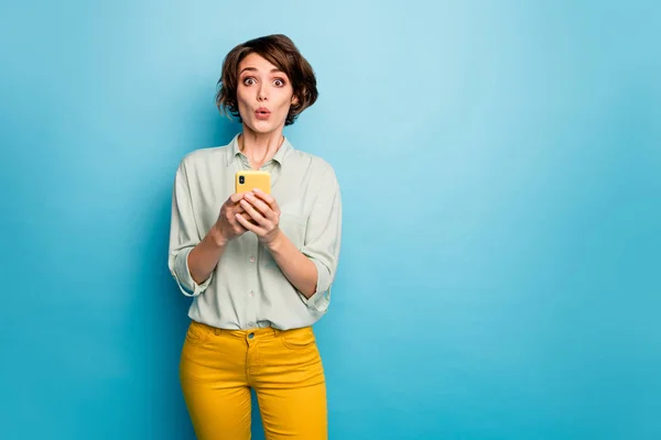 Foto van sprakeloze dame houden telefoon handen lees nieuwe blog opmerkingen niet geloven ogen doodsbang slijtage casual groen shirt geel broek geïsoleerde blauwe kleur achtergrond — Stockfoto