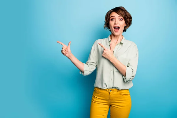 Foto de señora atractiva divertida que indica los dedos espacio vacío que aconseja precios bajos frescos compras ropa de boca abierta camisa verde casual pantalones amarillos aislados fondo de color azul —  Fotos de Stock