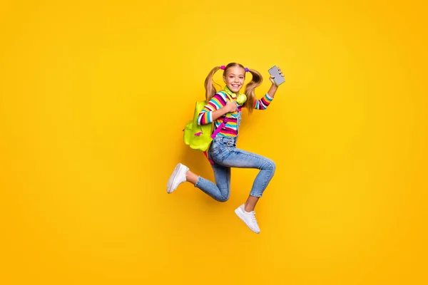 Full body profile side photo kid jump run fast lesson schoolgirl hold smartphone headset striped sweater denim jeans rucksack bag suspenders overalls isolated bright shine yellow color background — Φωτογραφία Αρχείου