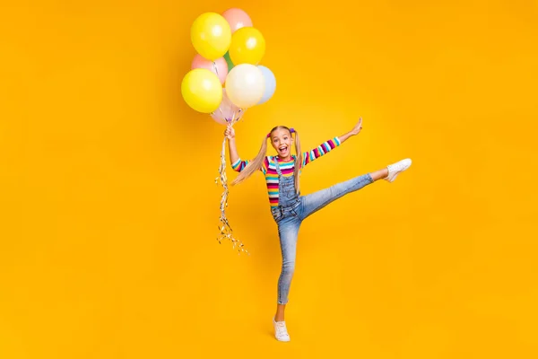 Full size photo of positive cheerful kid hold many helium baloons raise legs do gymnastics exercise wear denim outfit footwear isolated over yellow color background — Φωτογραφία Αρχείου