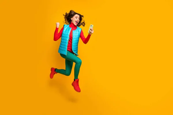 Teljes méretű fotó örömteli lány használata okostelefon kövesse a közösségi média ősz kedvezmény nyeremény lottó emelni öklöt sikoly ugrás futás kopás zöld kék piros nadrág cipő mellény elszigetelt sárga szín háttér — Stock Fotó
