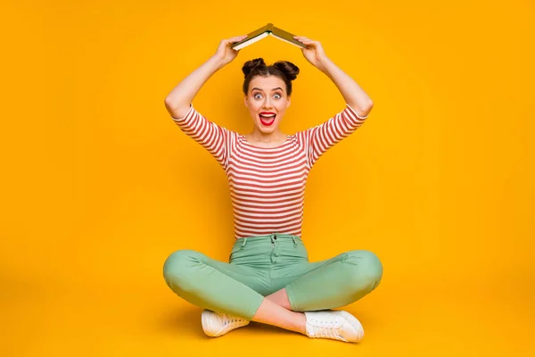 Full length photo of pretty lady sit floor hold book above head avoid do homework open mouth playful wear striped red white shirt green pants footwear isolated yellow color background — Stock Photo, Image