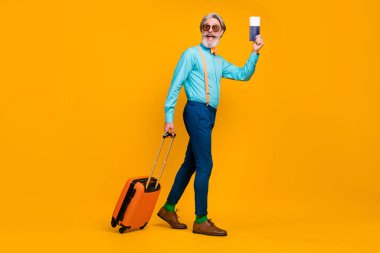Full size photo of cool grandpa hold tickets rolling suitcase walk registration table raise passport wear blue shirt suspenders bow tie pants boots socks isolated yellow color background clipart