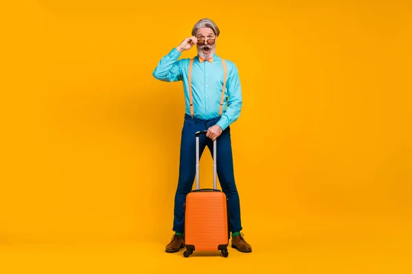 Полная задержка полета фото изумленный дедушка держит катящийся чемодан регистрации аэропорта носить очки подтяжки рубашки галстук-бабочка брюки носки изолированные желтый цвет фона — стоковое фото