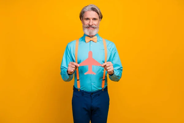 Foto de fresco abuelo elegante celebrar avión de papel recomendando elegir nueva aerolínea transporte desgaste camisa azul tirantes lazo pantalones aislados color amarillo fondo —  Fotos de Stock