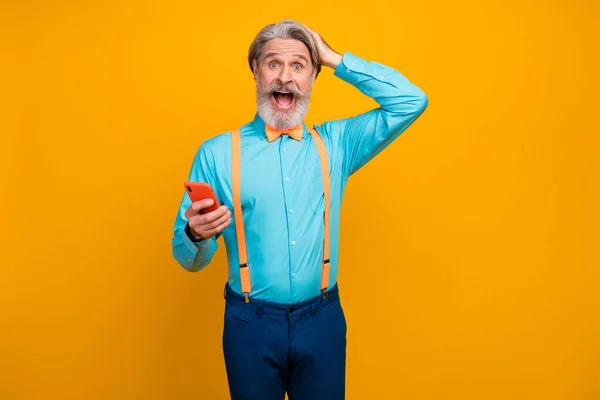 Foto de fresco impactado abuelo mantenga teléfono boca abierta sorprendido nuevo modelo de teléfono inteligente desgaste camisa azul tirantes lazo pantalones aislados color amarillo fondo —  Fotos de Stock