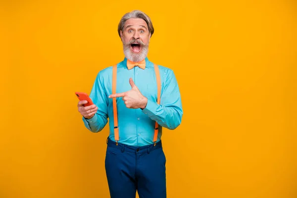 Foto do vovô legal segurar telefone direcionando tela de dedo boca aberta interface do dispositivo surpreso usar suspensórios camisa azul arco calça gravata isolado cor amarela fundo — Fotografia de Stock