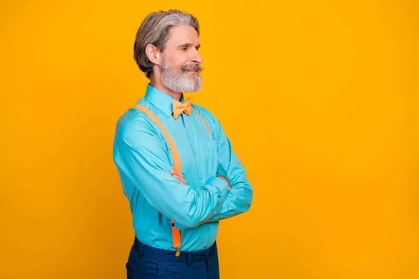 Profil foto av snygga coola kläder morfar gott humör händer korsade toothy leende ser tomt utrymme bära blå skjorta hängslen rosett byxor isolerad gul färg bakgrund — Stockfoto
