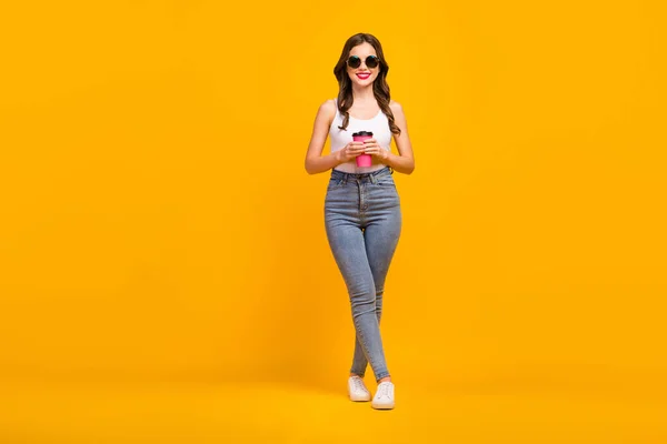 Full length photo of attractive cheerful lady good mood walk street drink hot takeaway coffee wear retro specs white tank-top jeans footwear isolated bright yellow color background — Stockfoto