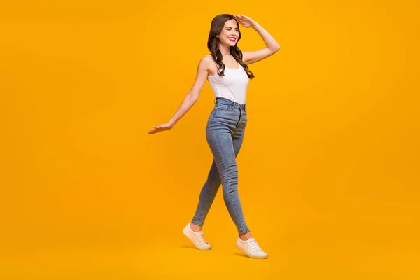 Full length profile photo of cheerful lady good mood look far away arm near forehead walk street wear white tank-top jeans footwear isolated bright yellow color background — Stockfoto