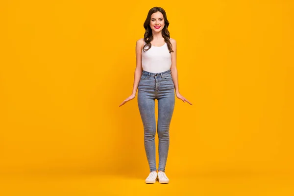 Full length photo of pretty shiny cheerful lady standing good mood slim figure shapes energetic person wear white tank-top jeans footwear isolated bright yellow color background — ストック写真