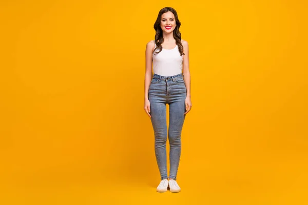 Full body photo of funny pretty shiny lady standing one place good mood slim figure shapes wear white tank-top jeans footwear isolated bright yellow color background — ストック写真