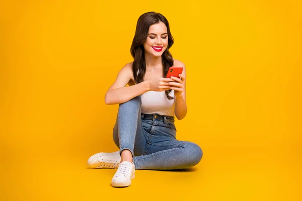Full length photo of funny lady sit floor cozy red lips holding telephone hands toothy smile wear white tank-top jeans gumshoes isolated bright yellow color background — Stockfoto