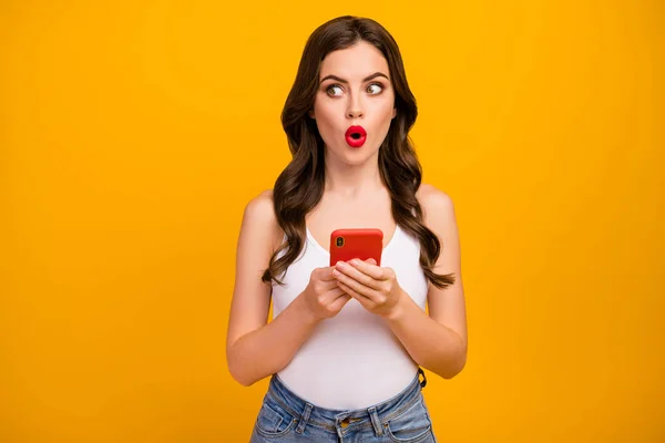 Photo of pretty shocked lady speechless hold telephone hands open mouth look side empty space cunning wear white tank-top jeans isolated bright yellow color background — Stock Fotó