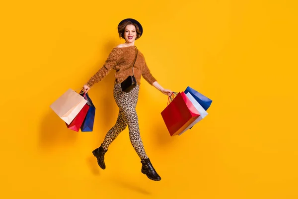 Teljes méretű fotó funky hölgy ugrás nagy örömteli szezon értékesítési bevásárló központ csomagok viselnek bolyhos pulóver leopárd nadrág csizma retro sapka válltáska elszigetelt sárga színű háttér — Stock Fotó