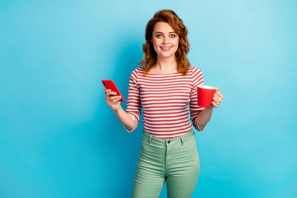 Портрет позитивної веселої жінки тримає чашку з латте використовують смс-повідомлення смартфона, друкуючи повідомлення про соціальну мережу, носити зелені штани, ізольовані на синьому кольорі — стокове фото