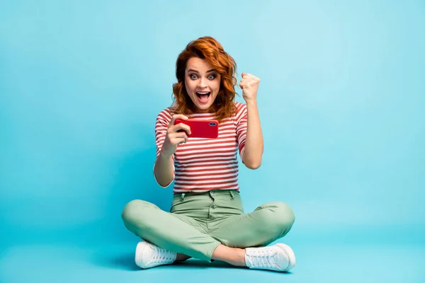 Full body photo delighted funky addicted woman sit legs crossed use cellphone read social media win lottery sms scream wow raise fists wear white green footwear isolated blue color background — ストック写真