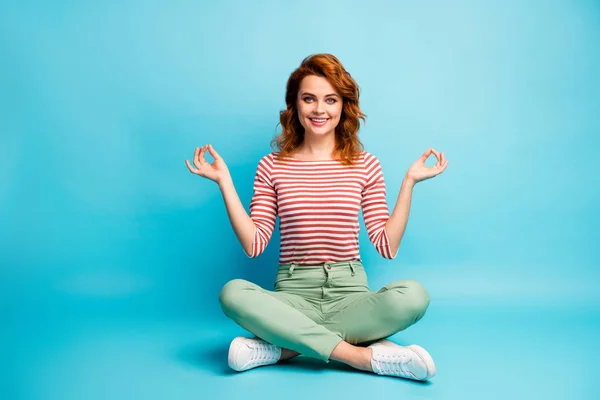 Full size photo of cheerful woman sit legs crossed enjoy peaceful calm sporty yoga exercise meditation show om sign wear white green footwear isolated over blue color background — Zdjęcie stockowe