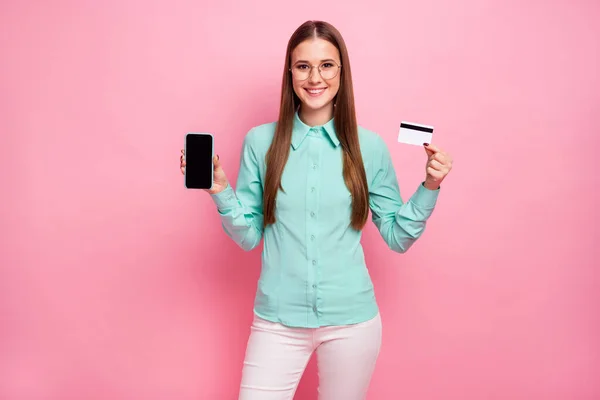 Retrato de niña alegre positivo celebrar banco tarjeta de crédito mostrar teléfono inteligente que compra en línea usar pantalones blancos pantalones formalwear turquesa aislado sobre fondo de color pastel —  Fotos de Stock