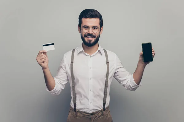 El hombre energético positivo sostiene la tarjeta de depósito, paga las facturas, ahorra los ingresos, usa el servicio de finanzas de teléfonos inteligentes, anuncia usar camisa blanca, pantalones marrones, pantalones aislados, fondo de color gris. —  Fotos de Stock