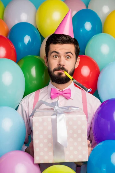 Κάθετη φωτογραφία του αστεία τύπος διαφορετικά χρώματα Noisemaker στο στόμα πάρτι γενεθλίων μεγάλο giftbox φορούν χάρτινο κώνο καπάκι ροζ πουκάμισο παπιγιόν τιράντες σε μπαλόνια δημιουργικό φόντο — Φωτογραφία Αρχείου