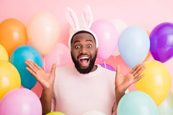 Foto di divertente ragazzo pelle scura inaspettate decorazioni a sorpresa festa pasquale umore festivo alzare le mani indossare maglione soffice coniglietto orecchie su palloncini colore rosa sfondo — Foto Stock