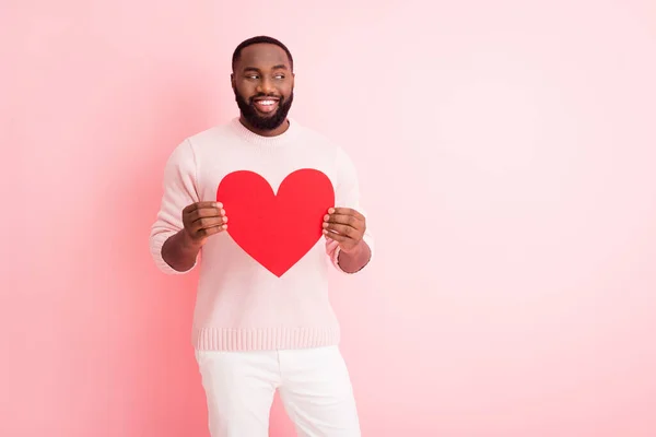 Foto de atractivo chico de piel oscura celebrar papel rojo corazón novio invitando a la novia romántica mirada lado espacio vacío desgaste suéter pantalones blancos aislados pastel rosa color fondo — Foto de Stock