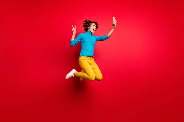 全长照片，疯狂时髦的黑发女孩跳过春秋假期，让自己的V字智能手机博客穿上蓝色毛衣黄色套装隔离明亮的红色背景 — 图库照片