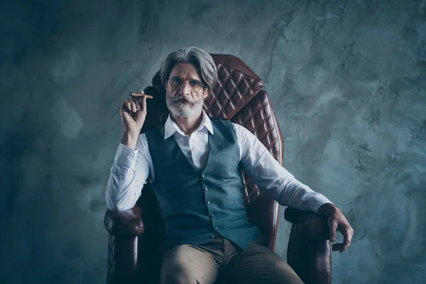Karizmatik yaşlı adam patron şirket sahibinin portresi. Kürsüye otur, puro tut. Gri arka planda izole edilmiş beyaz gömlek kahverengi pantolon giy. — Stok fotoğraf