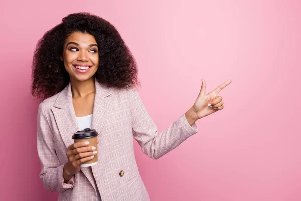 Positive afro american girl marketer representative hold take-out mug beverage point index finger present adverts promo wear checkered plaid jacket blazer isolated pastel pink color background