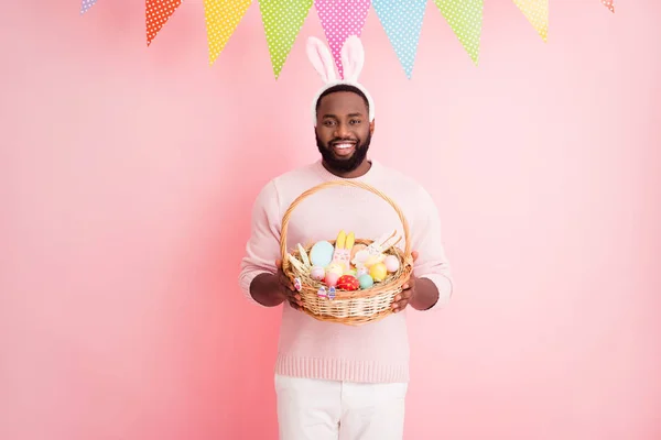 Photo of funny dark skin guy meet guests Wielkanoc party hold świąteczny kosz imbirowy chleb ciasteczka farba jaja nosić sweter spodnie króliczki uszy kolorowe flagi zawiesić różowy kolor tło — Zdjęcie stockowe