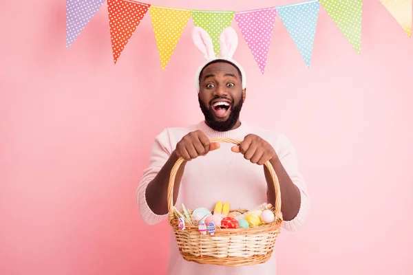 Foto di divertente scuro pelle ragazzo è venuto pasqua partito tenere festivo cesto zenzero pane dipinto uova biscotti indossare maglione coniglietto orecchie colorate bandiere appendere pastello colore rosa sfondo — Foto Stock