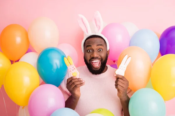 Photo of funny dark skin guy kolorowe dekoracje Wielkanoc trzymać dwie ładne ciasteczka imbirowe chleb nosić sweter królicze uszy na balony różowy kolor tło — Zdjęcie stockowe