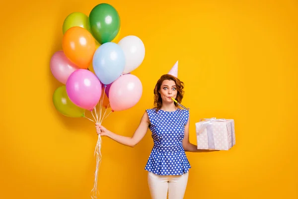 Foto der hübschen Dame halten viele bunte Luftballons große Geschenkbox Party Schlag Lärm Maker tragen Papier Kegelmütze Tupfen blaues Hemd weiße Hosen isoliert gelbe Farbe Hintergrund — Stockfoto