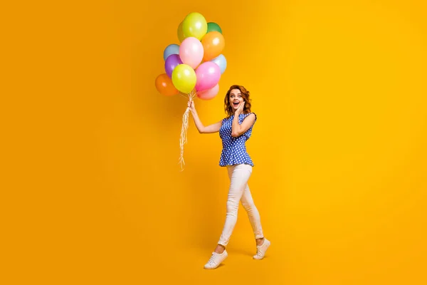 Foto de longitud completa de señora loca divertida celebrar muchos globos de colores caminar fiesta en movimiento evento de cumpleaños desgaste polka-dot azul camisa pantalones blancos zapatos aislados de color amarillo fondo —  Fotos de Stock