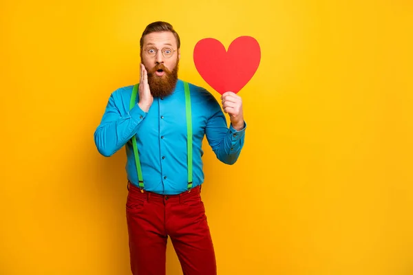 Foto de chico guapo sostener papel grande amantes del corazón día fecha invitación llena de alegría inesperada sorpresa desgaste azul camisa verde tirantes pantalones rojos aislados color amarillo fondo —  Fotos de Stock