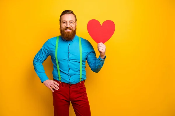 Foto de cara bonito segurar grande papel cartão postal coração chamando namorada romântico data amantes dia desgaste azul camisa verde suspensórios vermelho calças isoladas cor amarela fundo — Fotografia de Stock