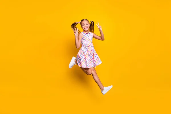 Full length photo of cheerful funny cute kid jump enjoy spring rest weekend holiday make v-signs wear pigtails ponytails gumshoes checkered skirt isolated bright yellow color background — Stok fotoğraf