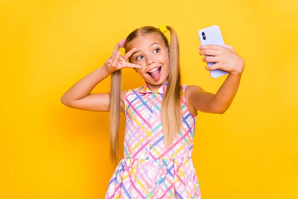 Foto de la señora bastante pequeña mantenga influencer teléfono hacer selfies blog mostrando símbolo de v-signo palo lengua fuera boca desgaste a cuadros vestido de verano aislado de color brillante fondo —  Fotos de Stock