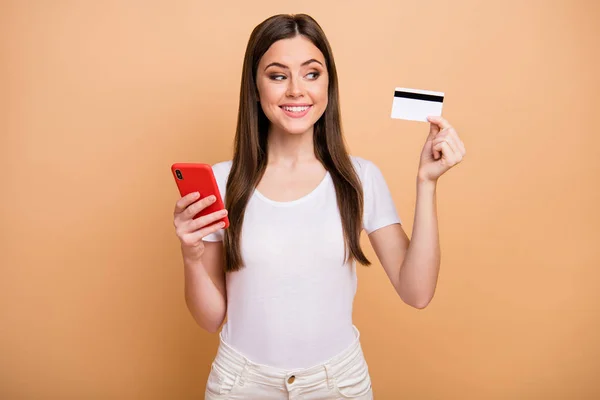 Retrato de niña alegre positiva mantenga mirada tarjeta de débito quieren compras en línea en la conexión del teléfono celular comprobar sus ganancias de ingresos en línea usar pantalones aislados color beige fondo —  Fotos de Stock