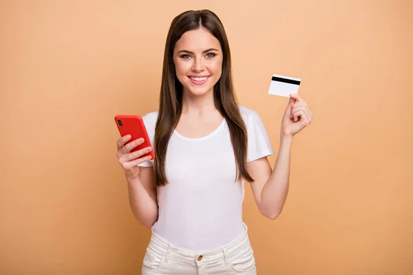 Retrato de chica positiva alegre depósito banco usuario celebrar teléfono celular uso tarjeta de crédito ahorros ganancias usar pantalones aislados sobre fondo de color beige —  Fotos de Stock