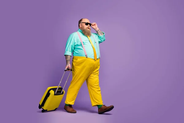 Вид профиля полноразмерного тела сбоку жизнерадостного зрелого парня, идущего с сумкой, трогательно прикасаясь к спецификациям вылета бюджетной авиакомпании, изолированной на ярком ярком светло-фиолетовом фоне — стоковое фото