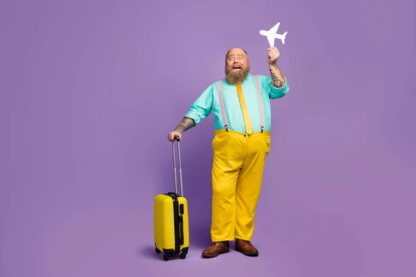 Full size photo excited man big abdomen hold papercard plane travel abroad weekend touch yellow suitcase wear shine turquoise shirt shoes pink suspenders pants isolated purple color background — ストック写真