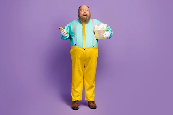 Full length photo excited overweight man use smartphone offer hold dream package prepare 14-february party wear teal shirt yellow pants pink suspenders isolated shine violet color background — Stok fotoğraf